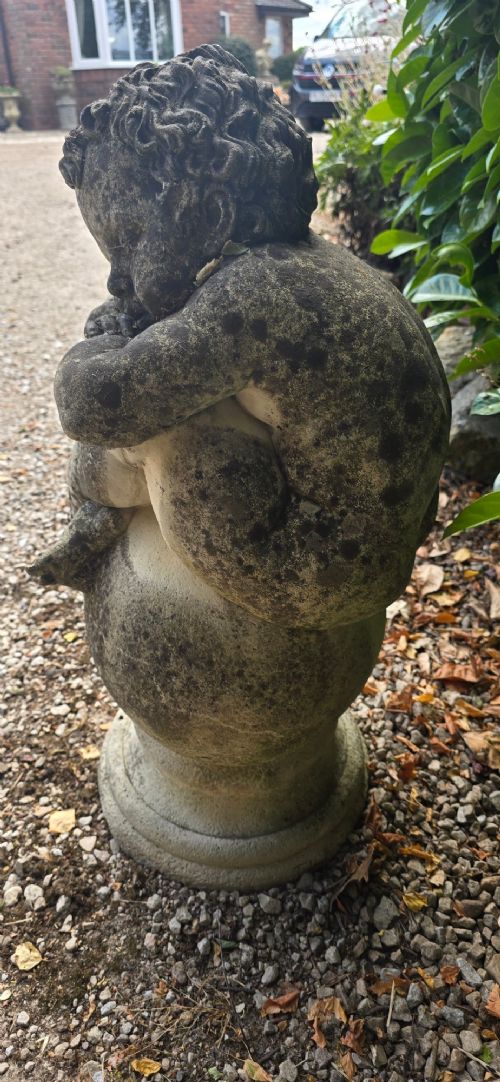 A weathered grey cast stone statue of a sleeping cherub resting its head on crossed arms atop a spherical base and moulded pedestal. View V