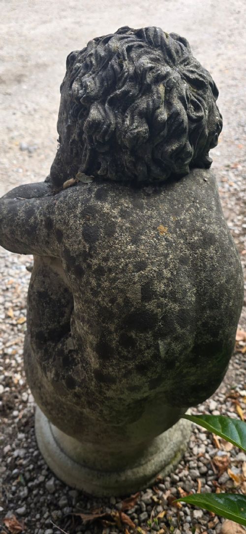 A weathered grey cast stone statue of a sleeping cherub resting its head on crossed arms atop a spherical base and moulded pedestal. View VI