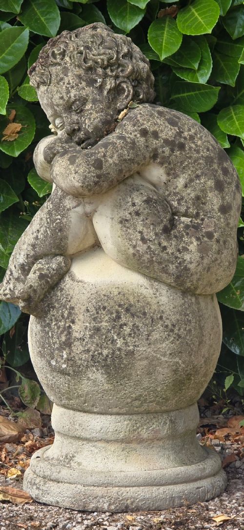 A weathered grey cast stone statue of a sleeping cherub resting its head on crossed arms atop a spherical base and moulded pedestal. View VIII