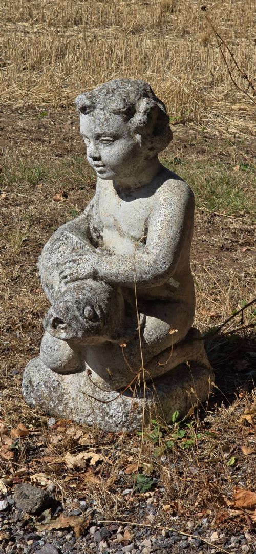 A weathered cast stone garden statue depicting a seated cherub holding a fish. View IV