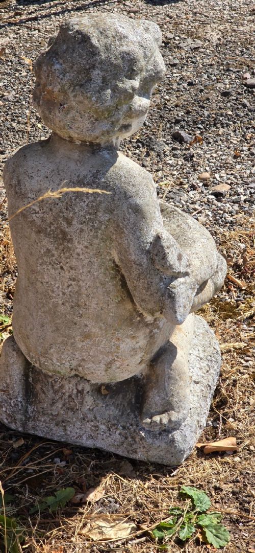 A weathered cast stone garden statue depicting a seated cherub holding a fish. View V