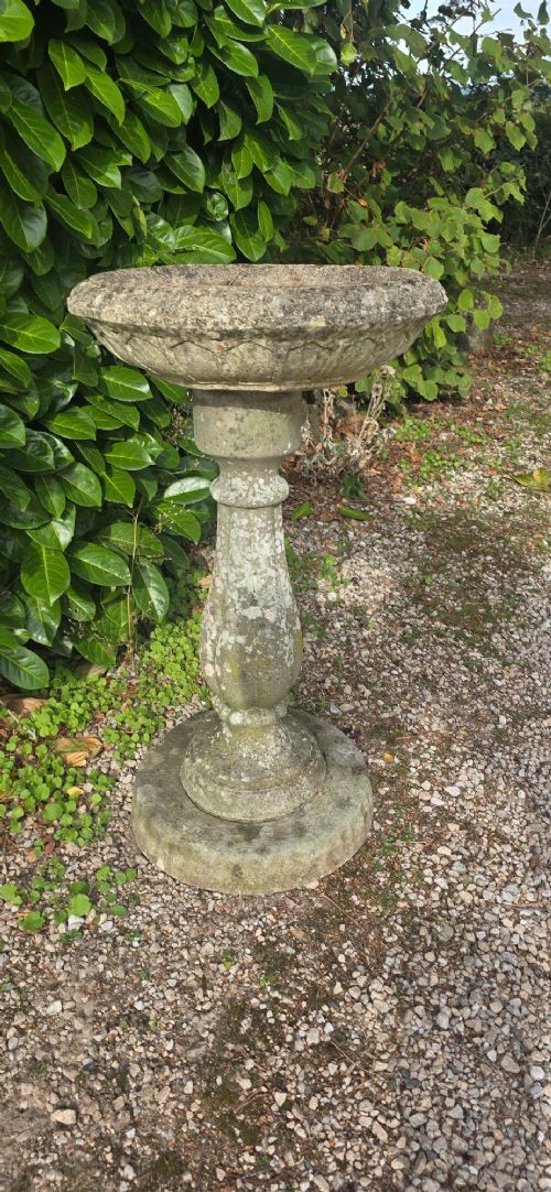 A weathered grey stone bird bath featuring a shallow circular bowl with carved rim detail on a baluster pedestal and round stepped base. View III