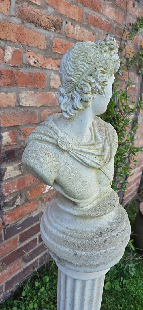 A light grey stone bust of Apollo Belvedere resting atop a matching fluted column plinth with visible outdoor weathering. View VI
