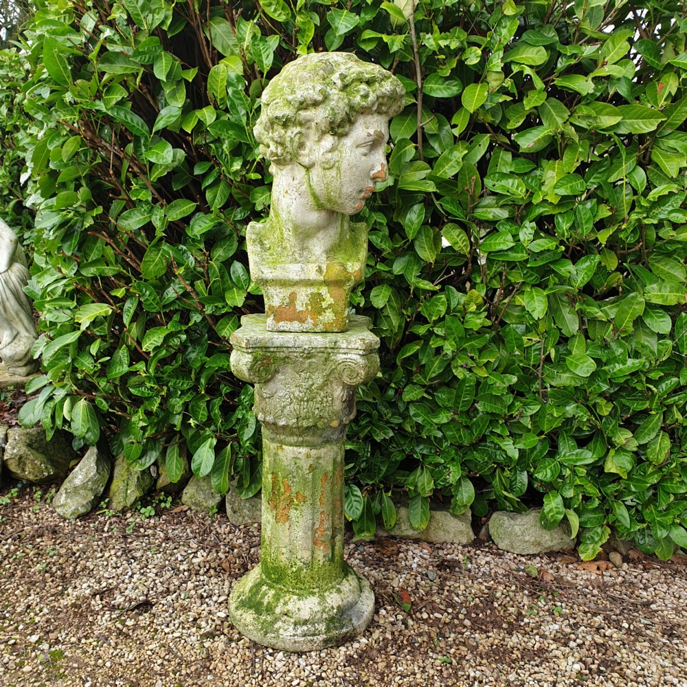 A classical stone bust of a male figure resting atop a fluted stone column pedestal with visible green lichen patination.