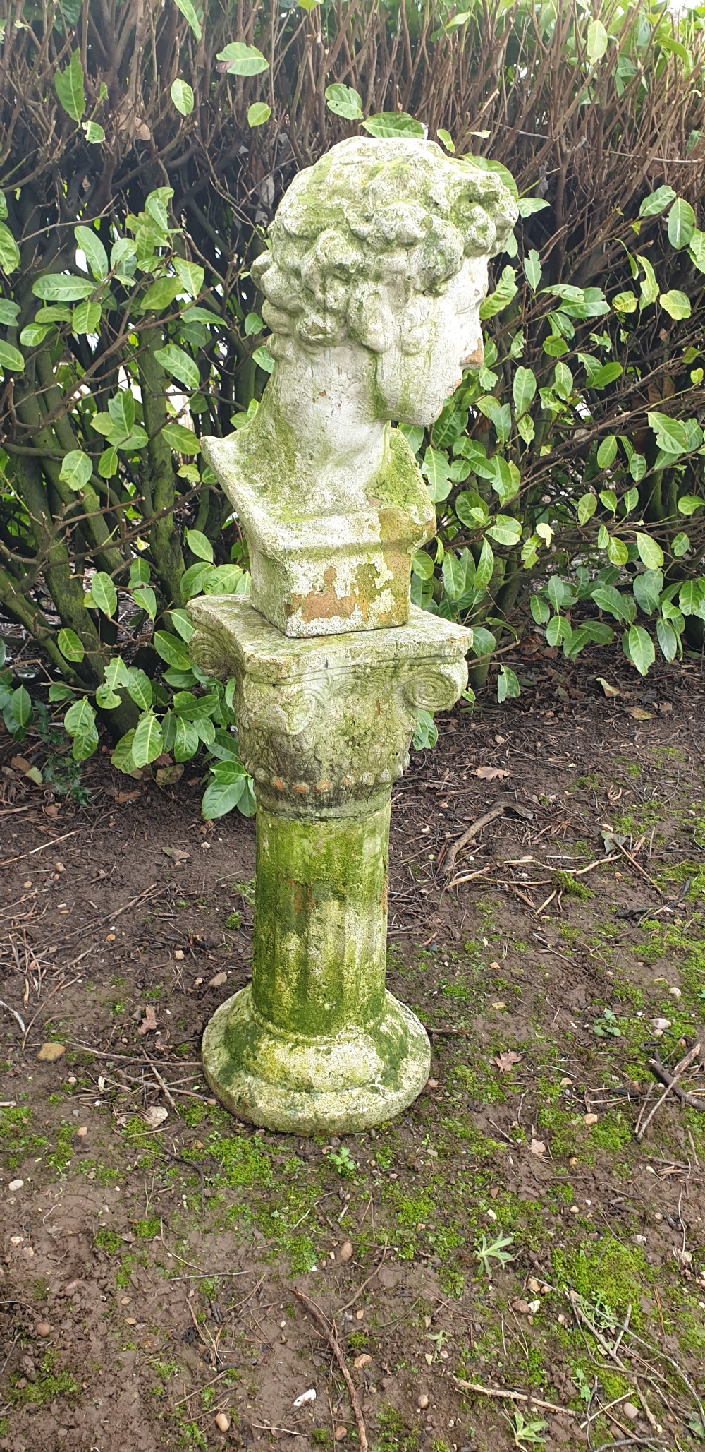 A classical stone bust of a male figure resting atop a fluted stone column pedestal with visible green lichen patination. View III