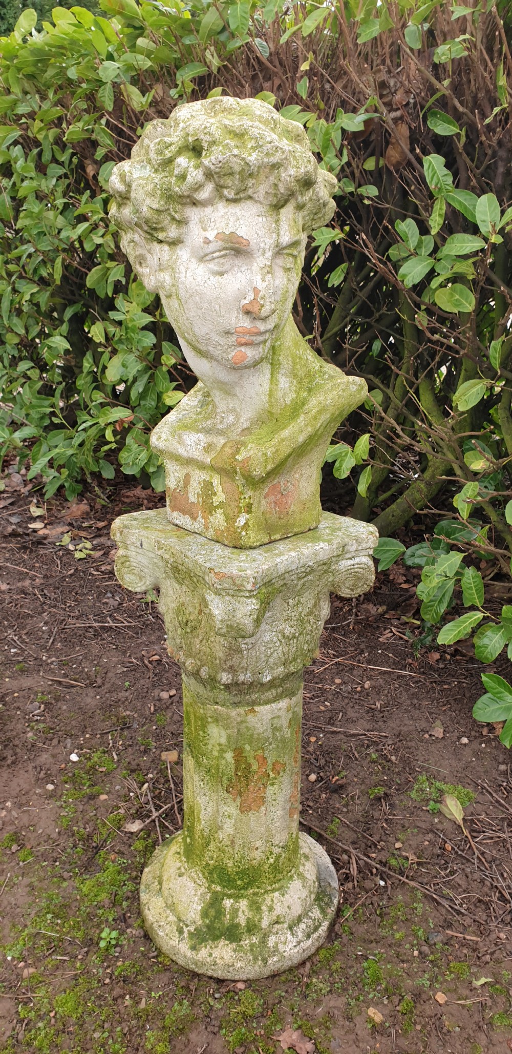 A classical stone bust of a male figure resting atop a fluted stone column pedestal with visible green lichen patination. View V