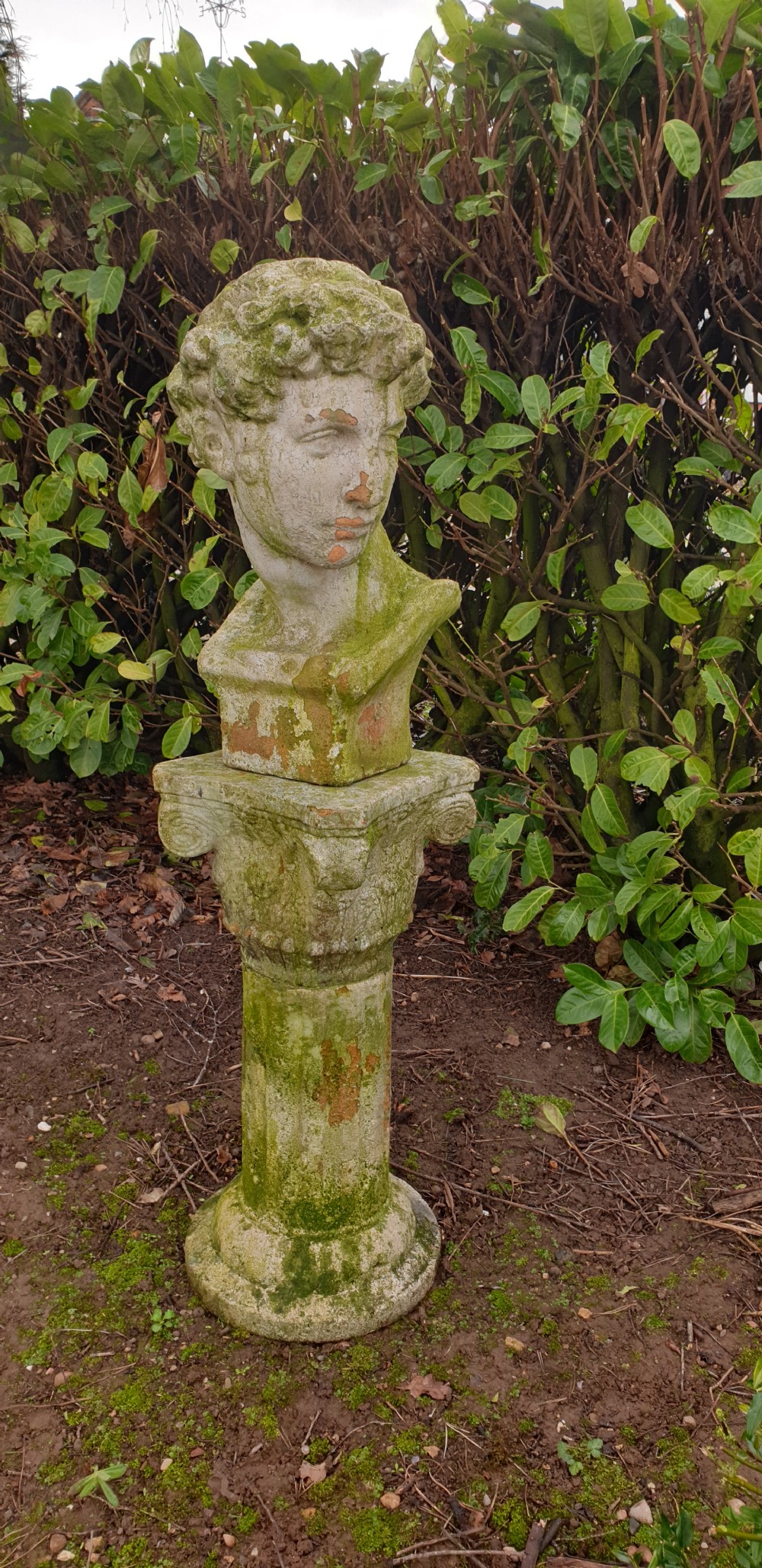 A classical stone bust of a male figure resting atop a fluted stone column pedestal with visible green lichen patination. View VI