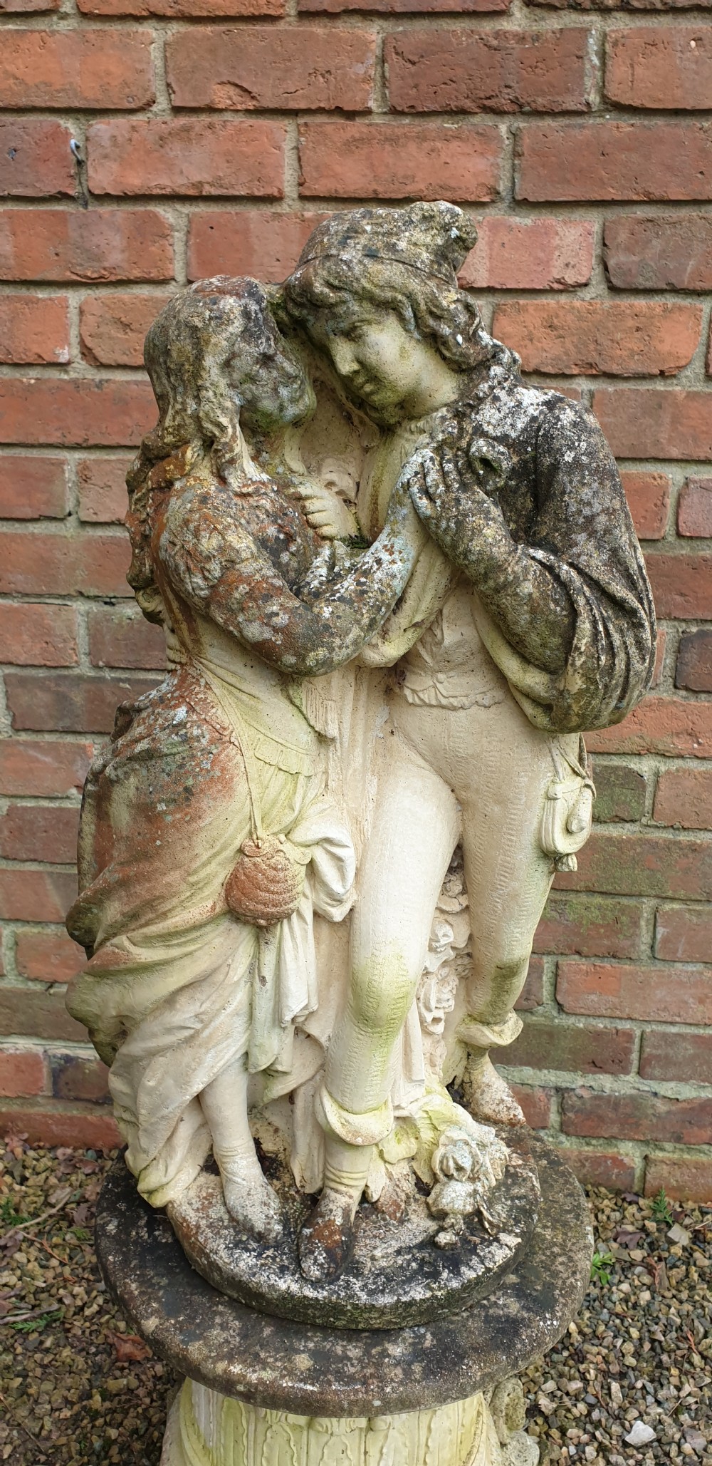 A weathered cast stone garden sculpture depicting a male and female figure embracing, positioned atop a carved pedestal. View II