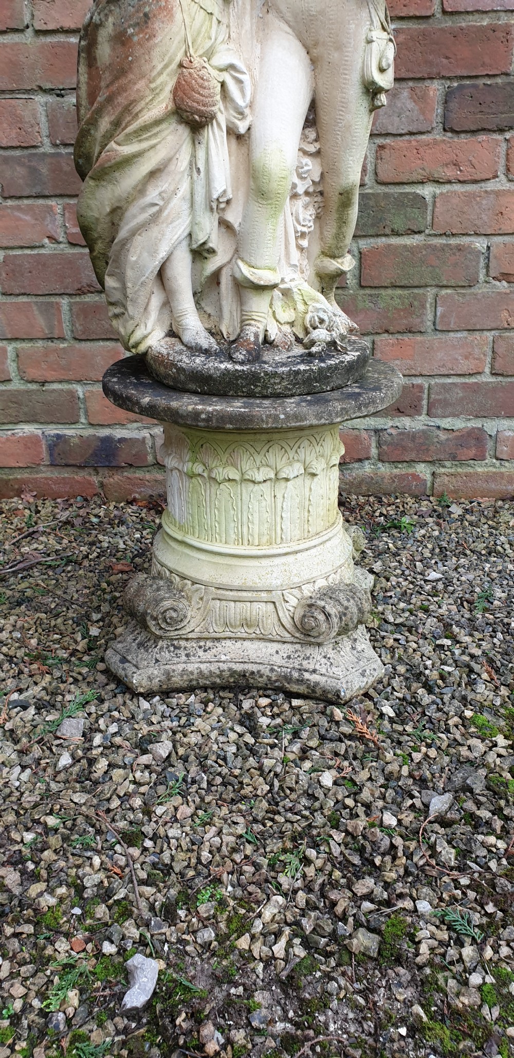 A weathered cast stone garden sculpture depicting a male and female figure embracing, positioned atop a carved pedestal. View III