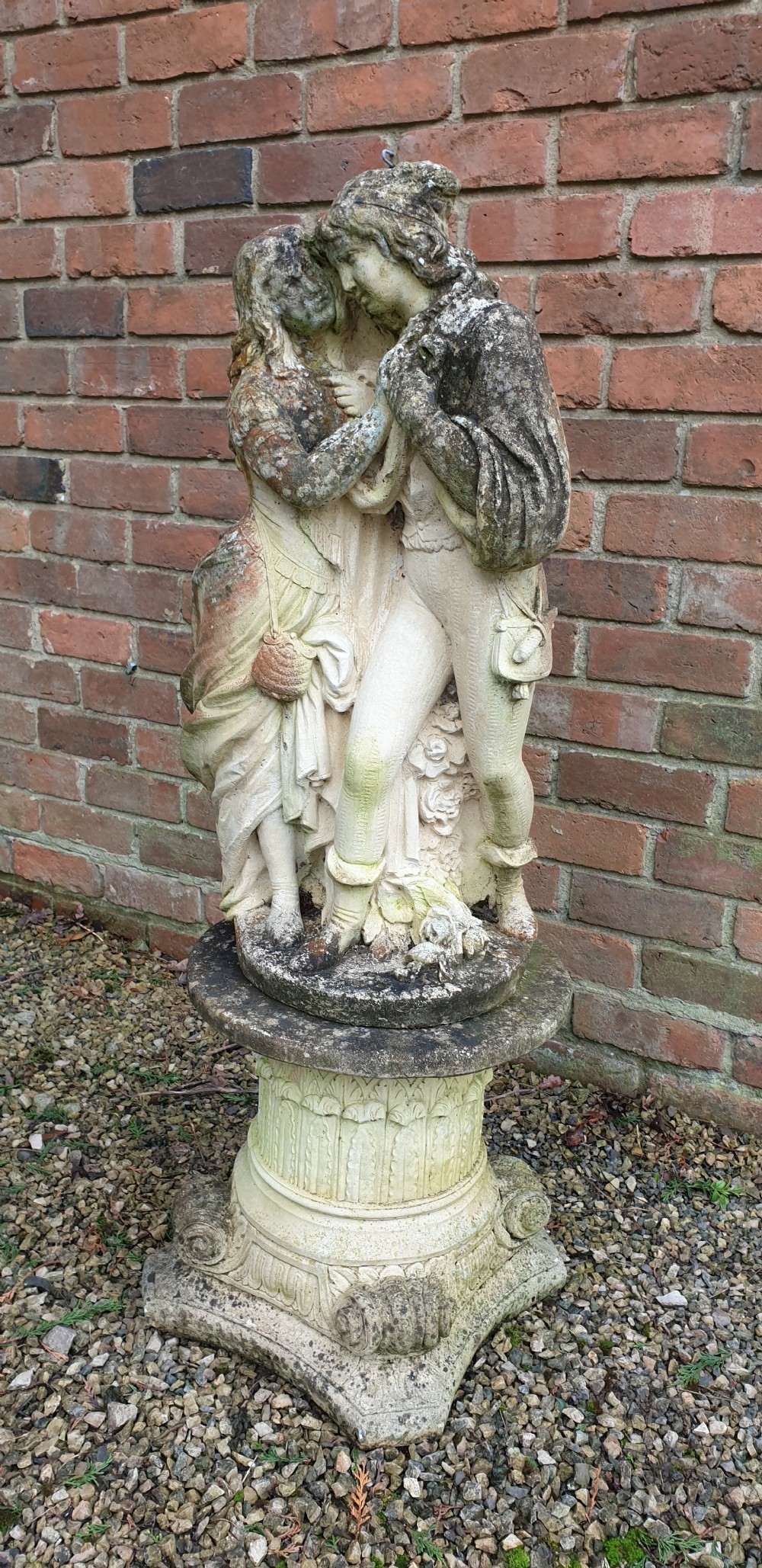 A weathered cast stone garden sculpture depicting a male and female figure embracing, positioned atop a carved pedestal. View IV