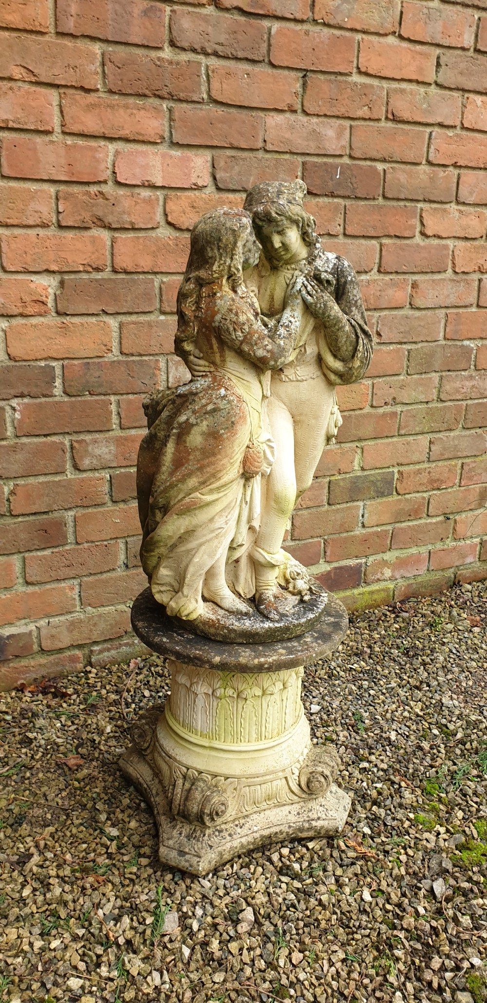 A weathered cast stone garden sculpture depicting a male and female figure embracing, positioned atop a carved pedestal. View V