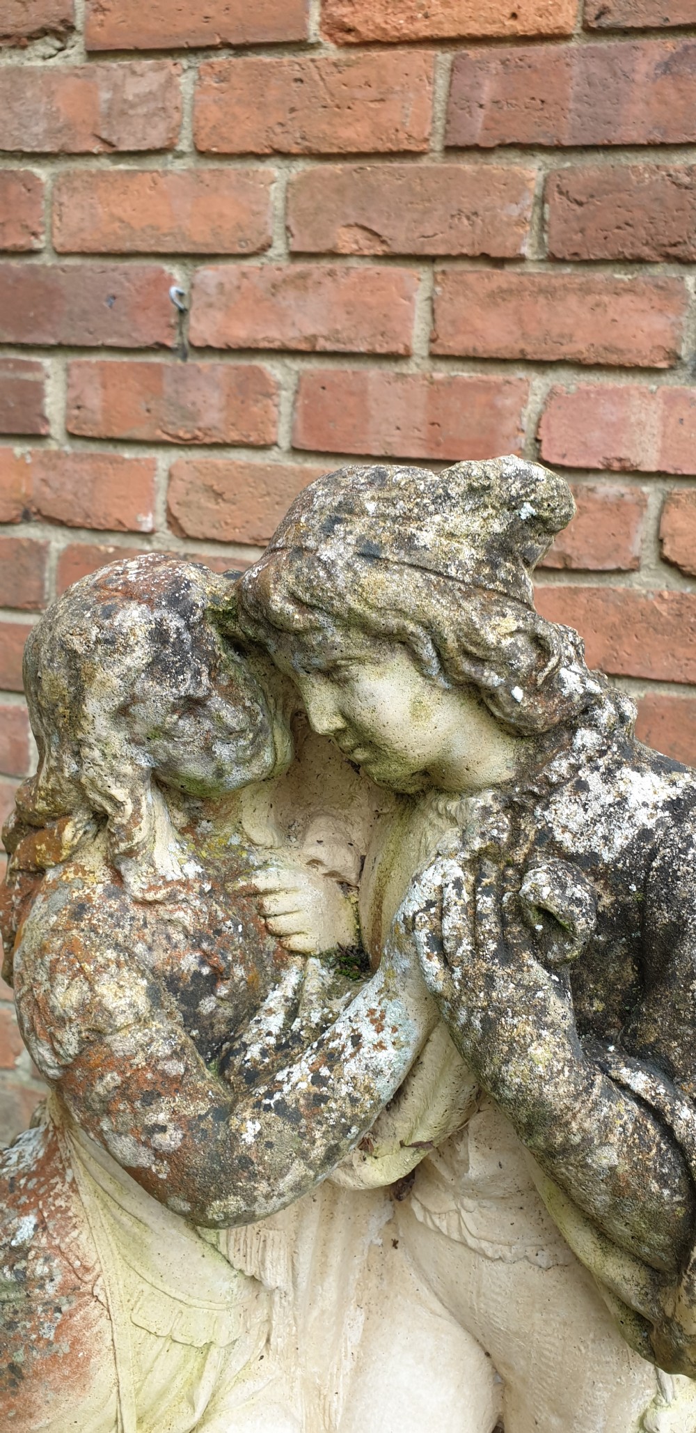 A weathered cast stone garden sculpture depicting a male and female figure embracing, positioned atop a carved pedestal. View VI