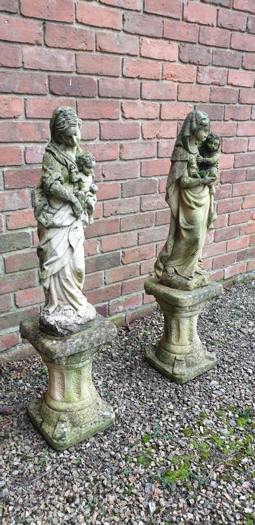 A pair of weathered grey stone statues depicting the Madonna and Child, each mounted on a square tapered plinth base. View VII