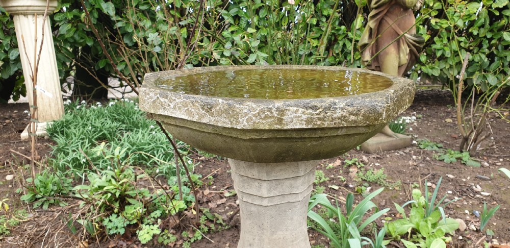 A weathered grey cast stone bird bath featuring an octagonal basin resting upon a tapered faceted pedestal base. View III