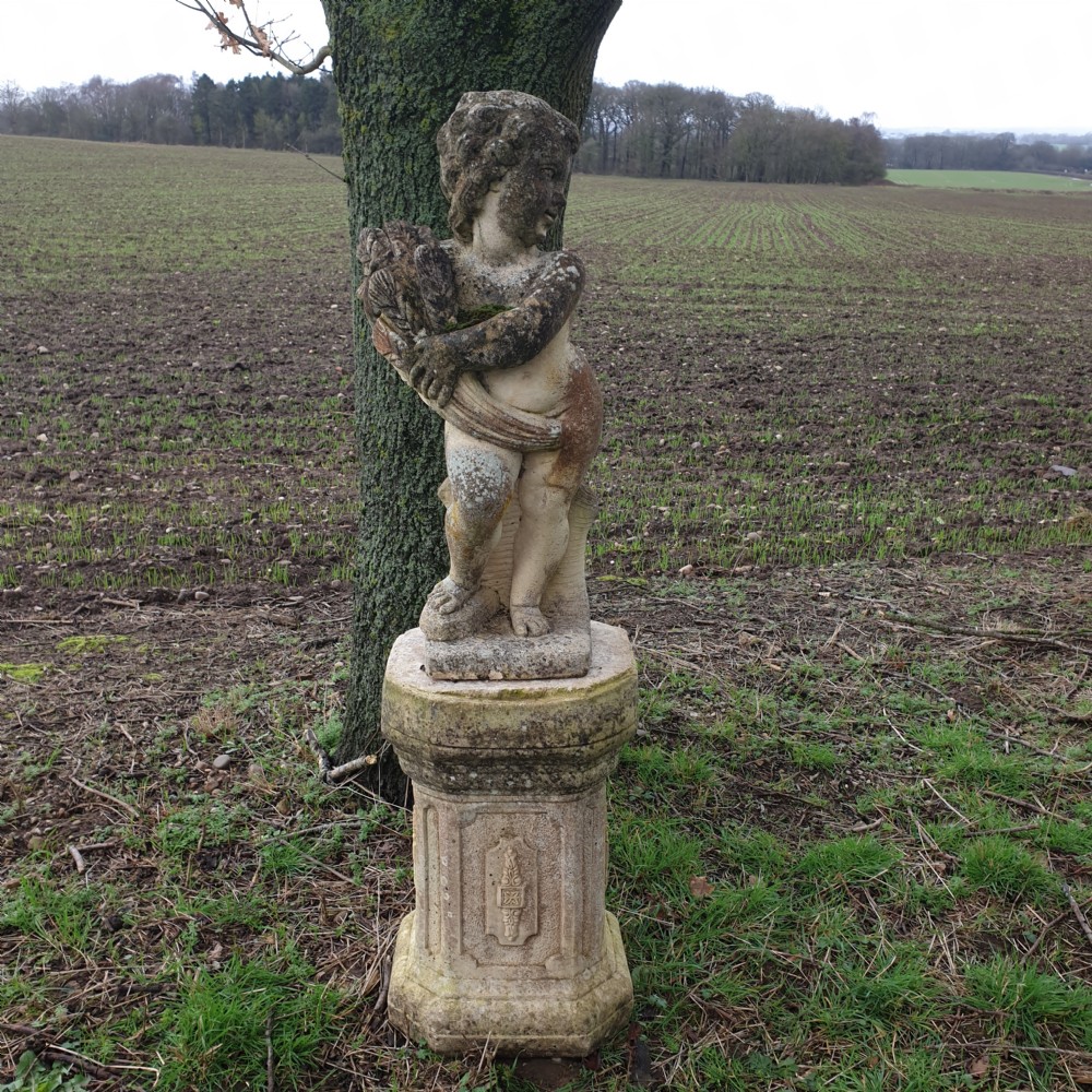 A weathered stone garden statue of a cherub holding a sheaf of grain, positioned atop an ornamental moulded stone pedestal.