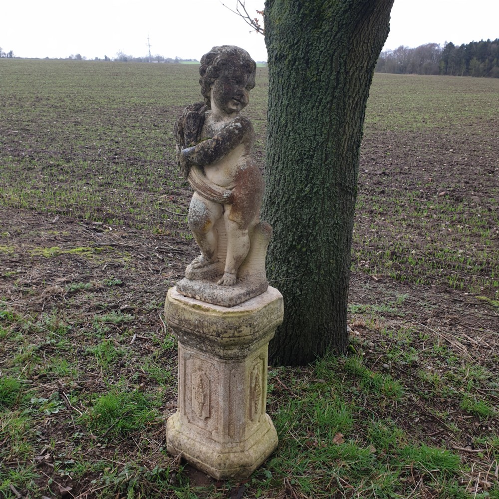 A weathered stone garden statue of a cherub holding a sheaf of grain, positioned atop an ornamental moulded stone pedestal. View II
