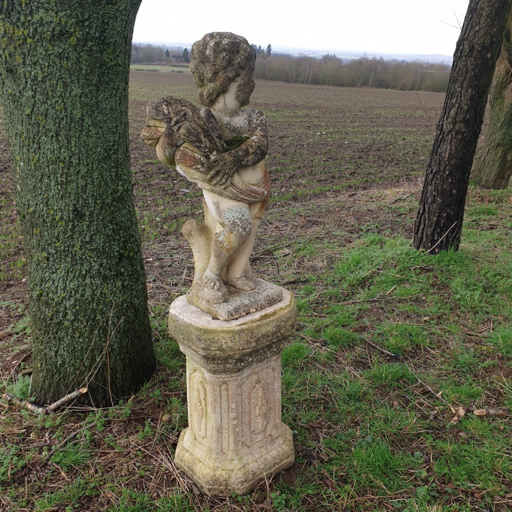 A weathered stone garden statue of a cherub holding a sheaf of grain, positioned atop an ornamental moulded stone pedestal. View III