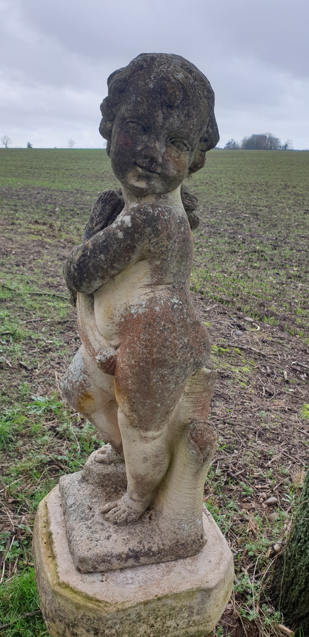 A weathered stone garden statue of a cherub holding a sheaf of grain, positioned atop an ornamental moulded stone pedestal. View VI
