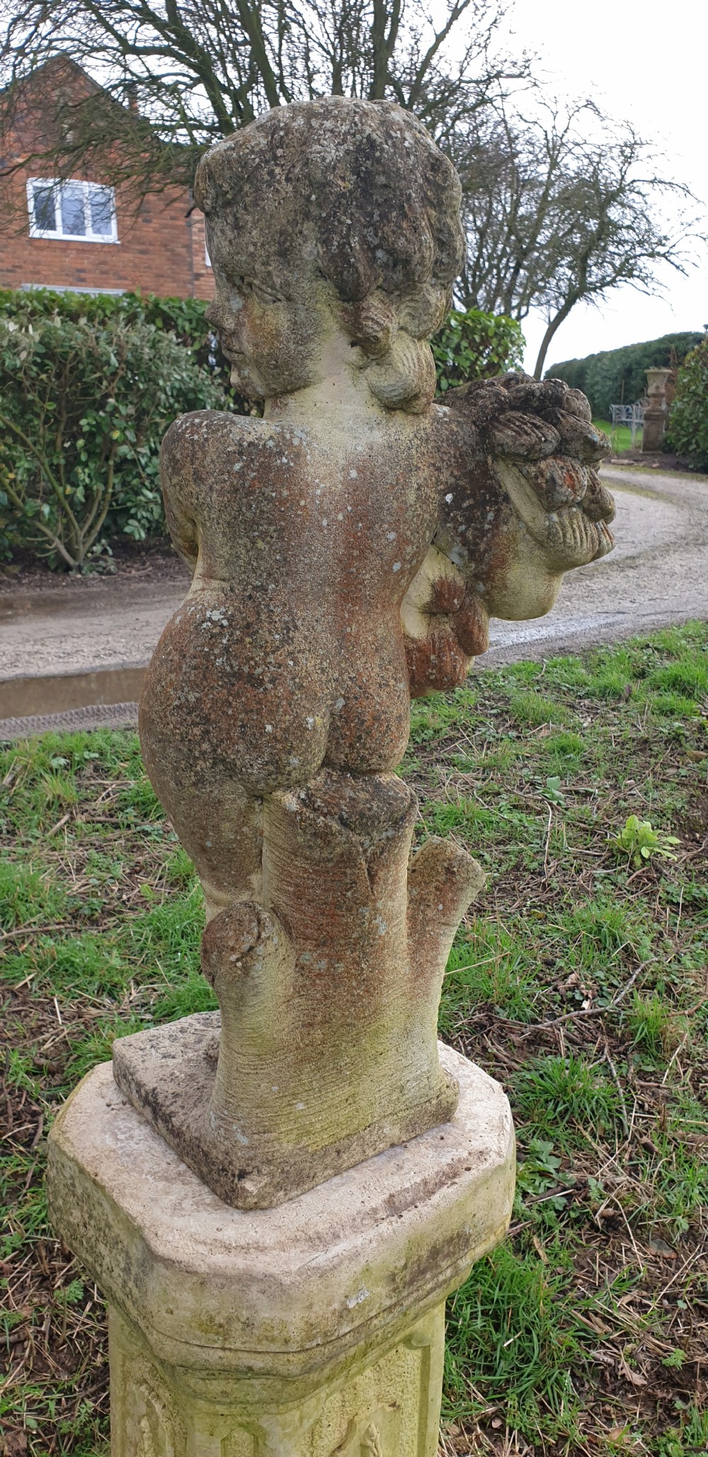 A weathered stone garden statue of a cherub holding a sheaf of grain, positioned atop an ornamental moulded stone pedestal. View VII