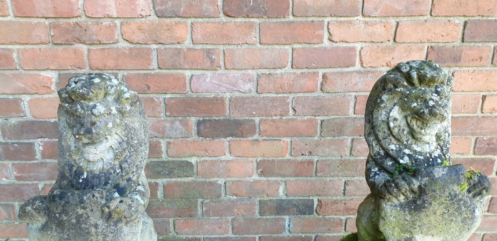 A pair of weathered grey stone seated lion statues holding shields, mounted on relief-carved rectangular plinths. View VI