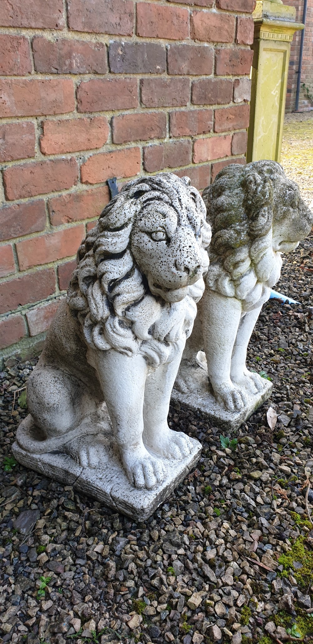 A pair of weathered off-white reconstituted stone lion statues rendered in a seated pose atop integral rectangular bases. View VI