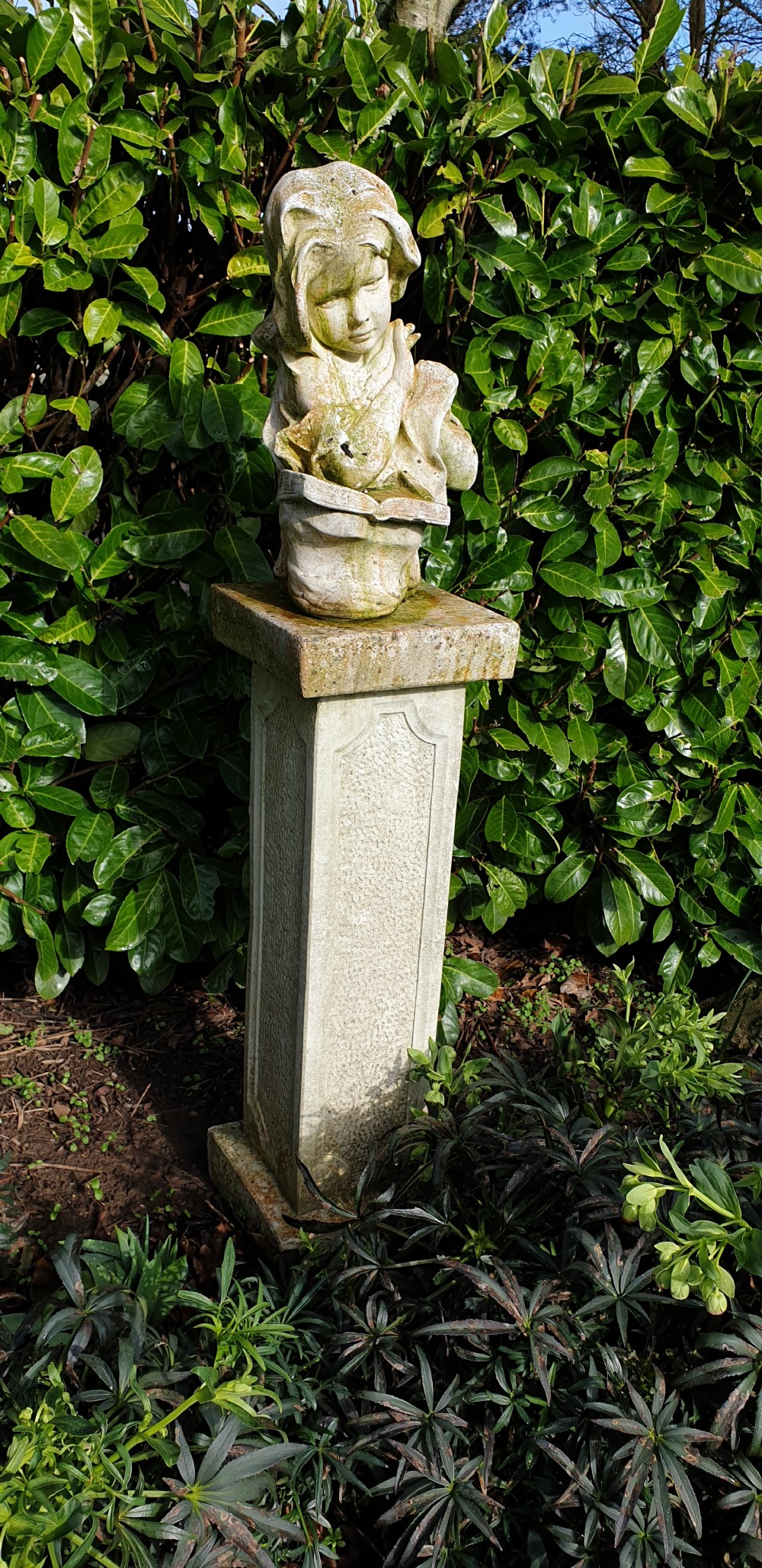 A weathered cast stone bust depicting a young child reading a book set upon a tall square pedestal plinth. View II