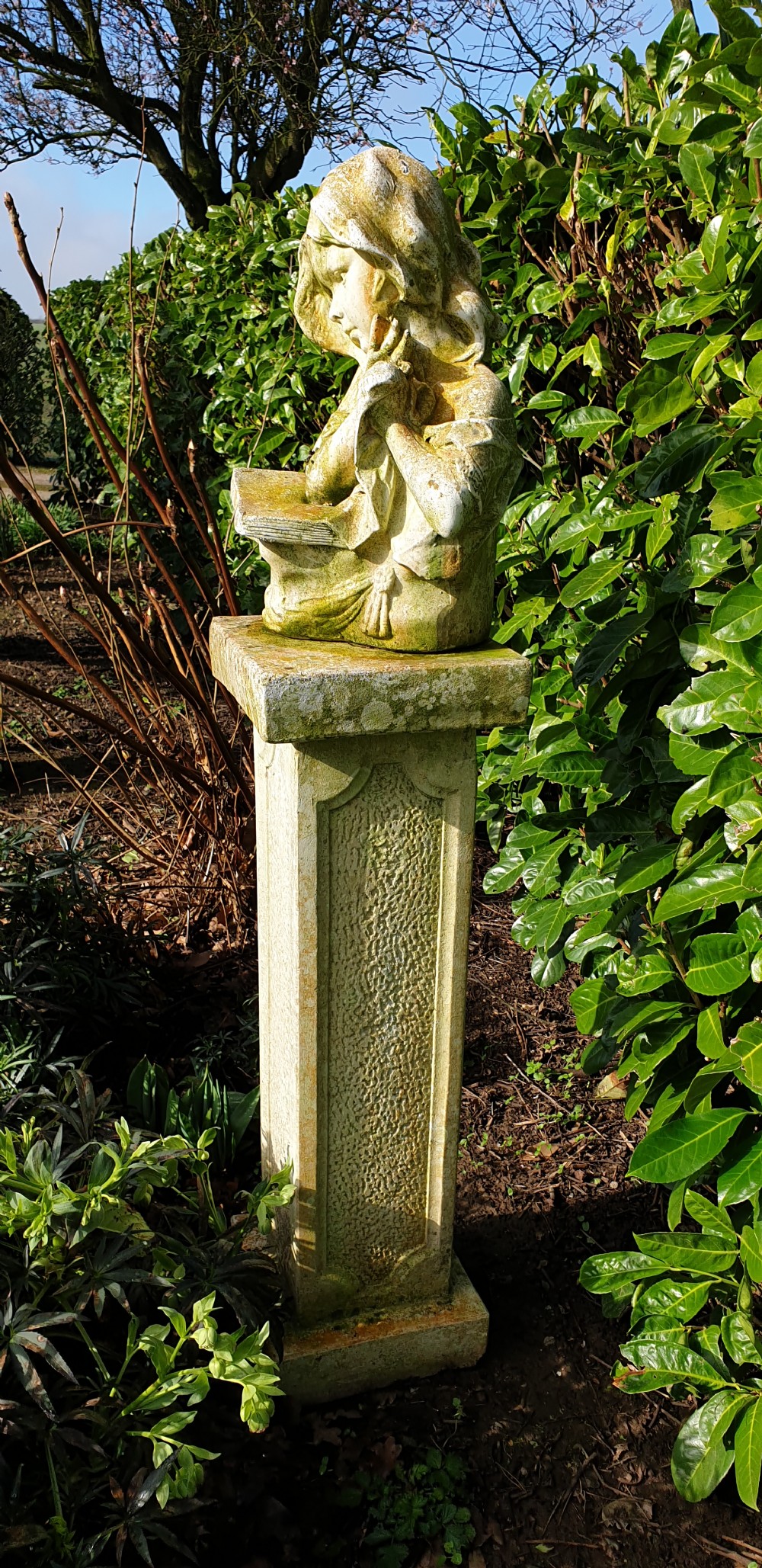 A weathered cast stone bust depicting a young child reading a book set upon a tall square pedestal plinth. View IV