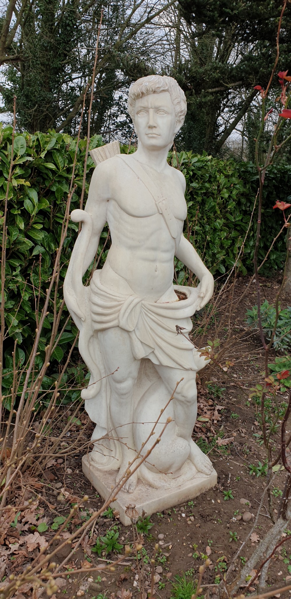 A life-sized off-white stone statue depicting a draped classical male figure holding a bow with a quiver on his back, set on a square plinth. View II