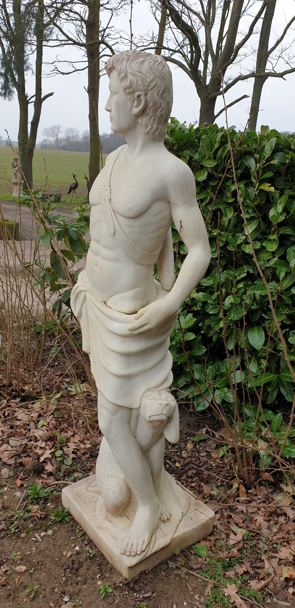 A life-sized off-white stone statue depicting a draped classical male figure holding a bow with a quiver on his back, set on a square plinth. View III