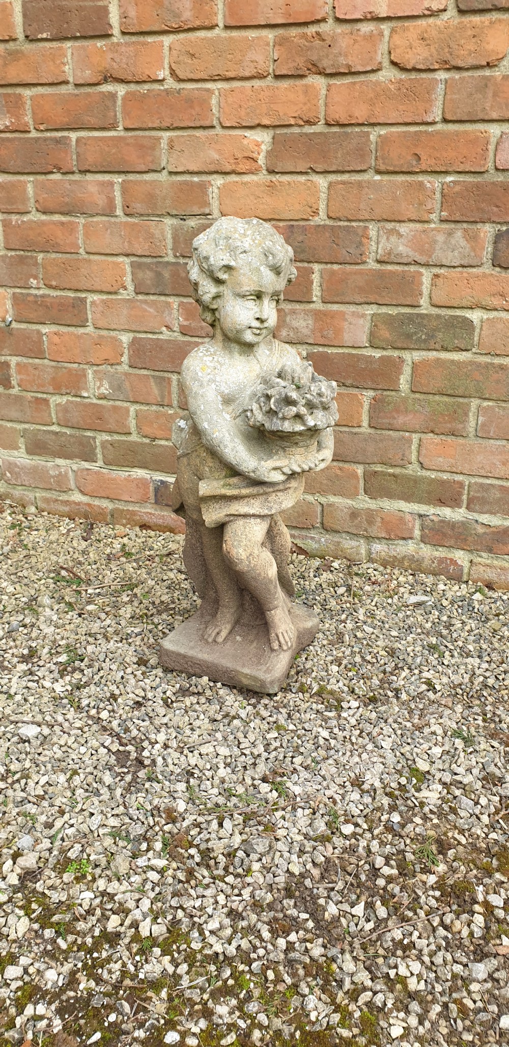 A weathered grey stone garden statue depicting a standing cherub holding a basket of flowers on an integral square plinth.