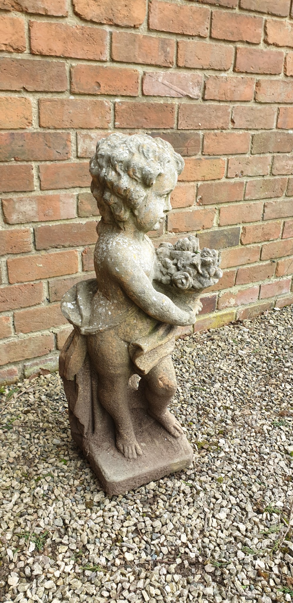 A weathered grey stone garden statue depicting a standing cherub holding a basket of flowers on an integral square plinth. View II