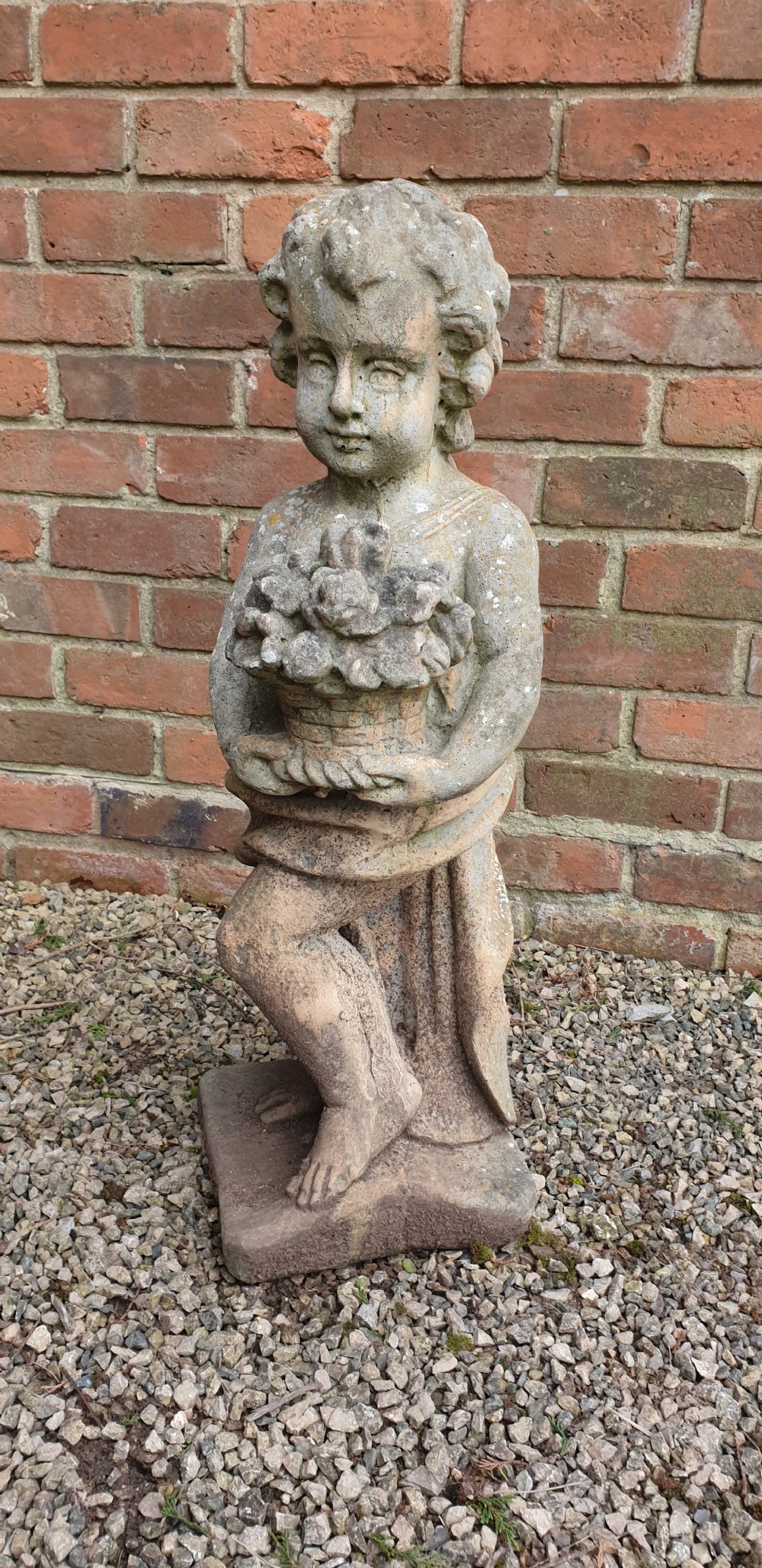 A weathered grey stone garden statue depicting a standing cherub holding a basket of flowers on an integral square plinth. View III