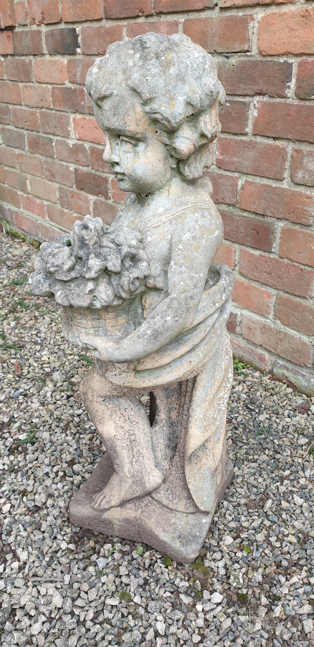 A weathered grey stone garden statue depicting a standing cherub holding a basket of flowers on an integral square plinth. View IV