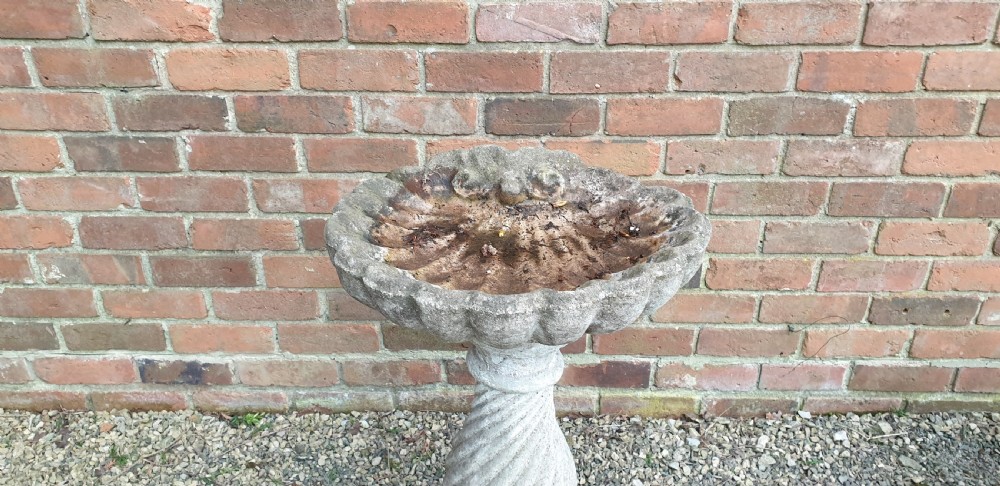 A weathered grey stone bird bath featuring a scalloped shell-shaped top basin set upon a spiral twisted pedestal column and square base. View V