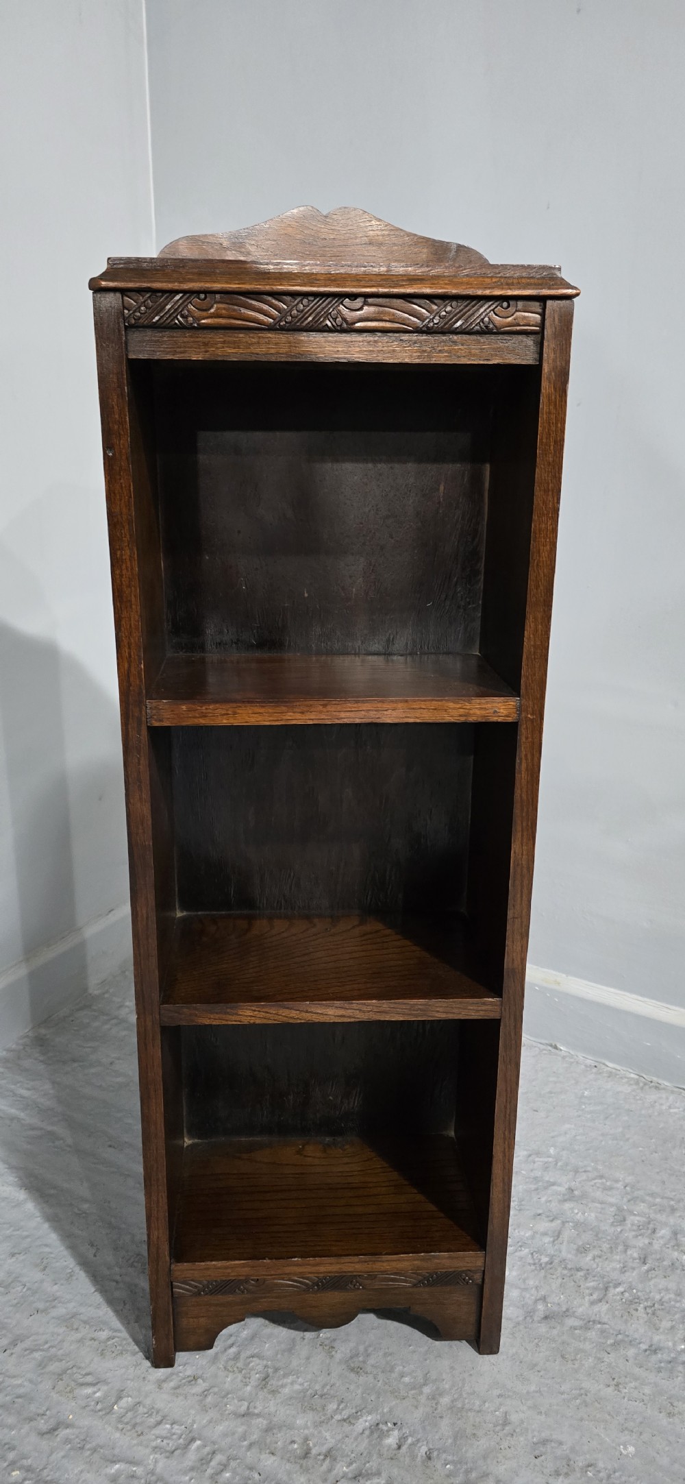 A narrow dark oak open bookcase featuring three fixed shelves, a crest gallery top, and a carved decorative frieze. View V