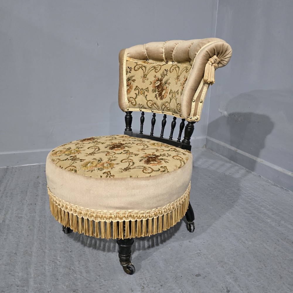 An Edwardian boudoir chair with gold floral upholstery, deep fringe trim around the circular base, turned spindles, and rolled velvet backrest on castors.