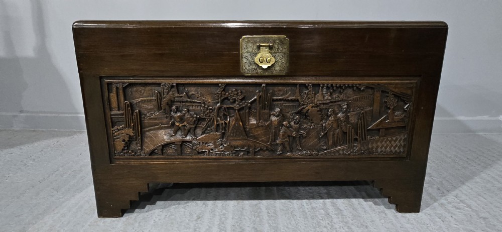 A dark brown camphor wood trunk featuring detailed relief carvings of figures and buildings on the front panel and a decorative brass latch plate.