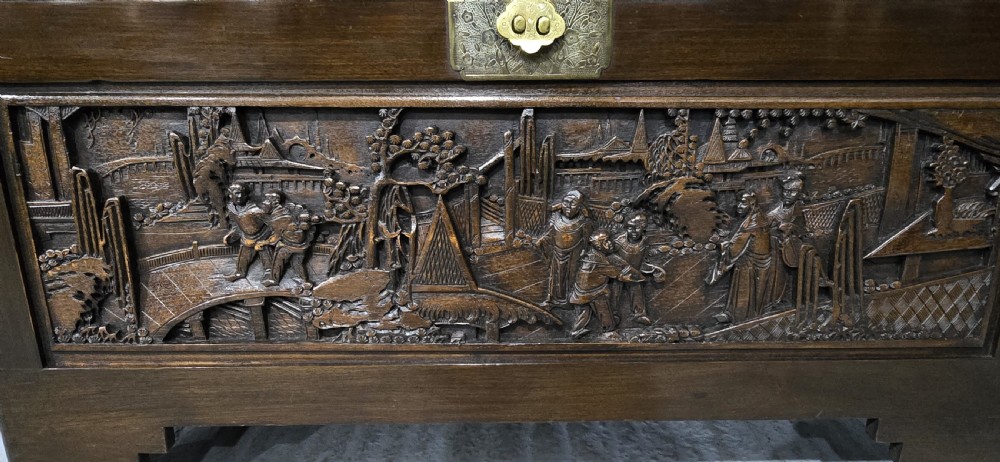 A dark brown camphor wood trunk featuring detailed relief carvings of figures and buildings on the front panel and a decorative brass latch plate. View II