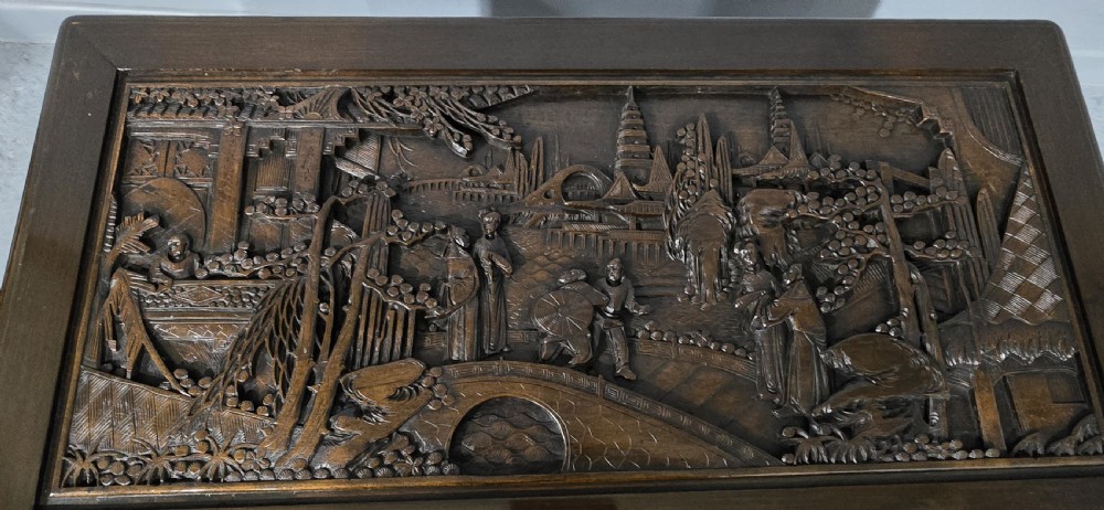 A dark brown camphor wood trunk featuring detailed relief carvings of figures and buildings on the front panel and a decorative brass latch plate. View III