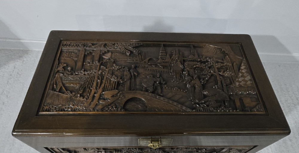 A dark brown camphor wood trunk featuring detailed relief carvings of figures and buildings on the front panel and a decorative brass latch plate. View V