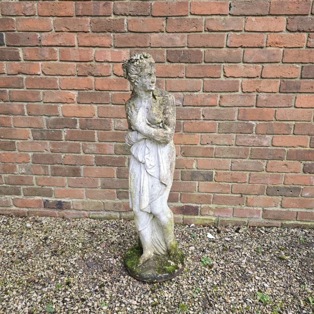 A weathered reconstituted stone statue depicting a draped female figure standing on an integrated circular base.