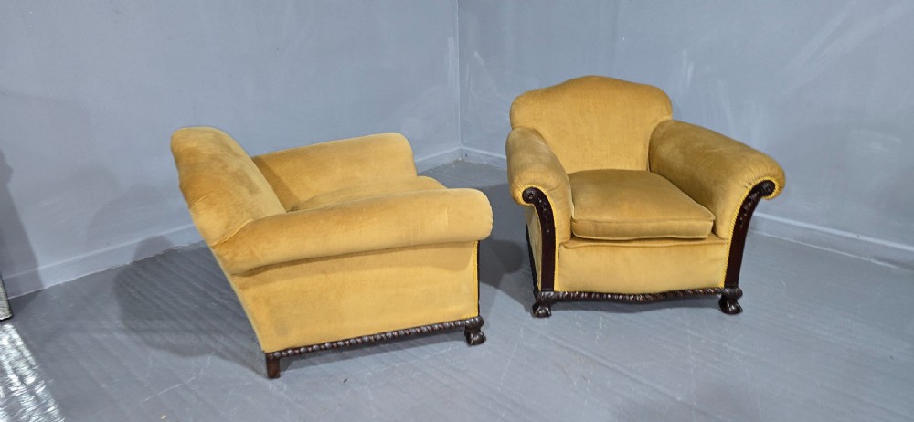 A pair of yellow velvet upholstered club chairs featuring carved dark timber trim along the front arms and base. View VI