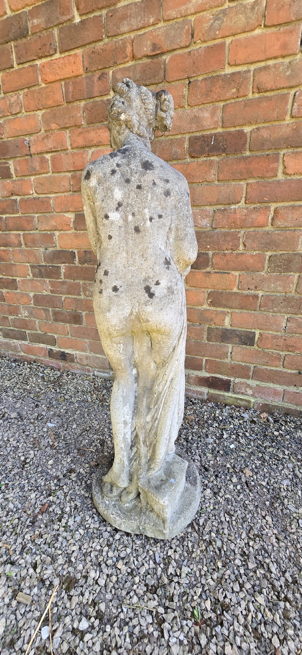 A weathered cast stone garden statue depicting a classical female figure draped in fabric standing on a round integral base. View II
