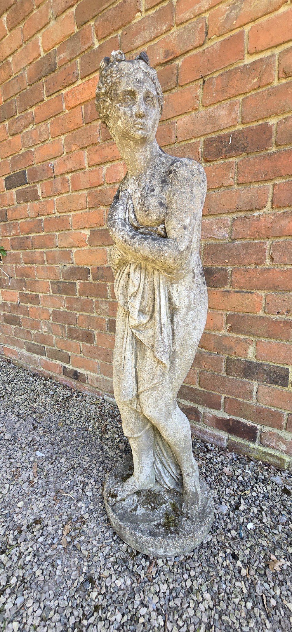 A weathered cast stone garden statue depicting a classical female figure draped in fabric standing on a round integral base. View V