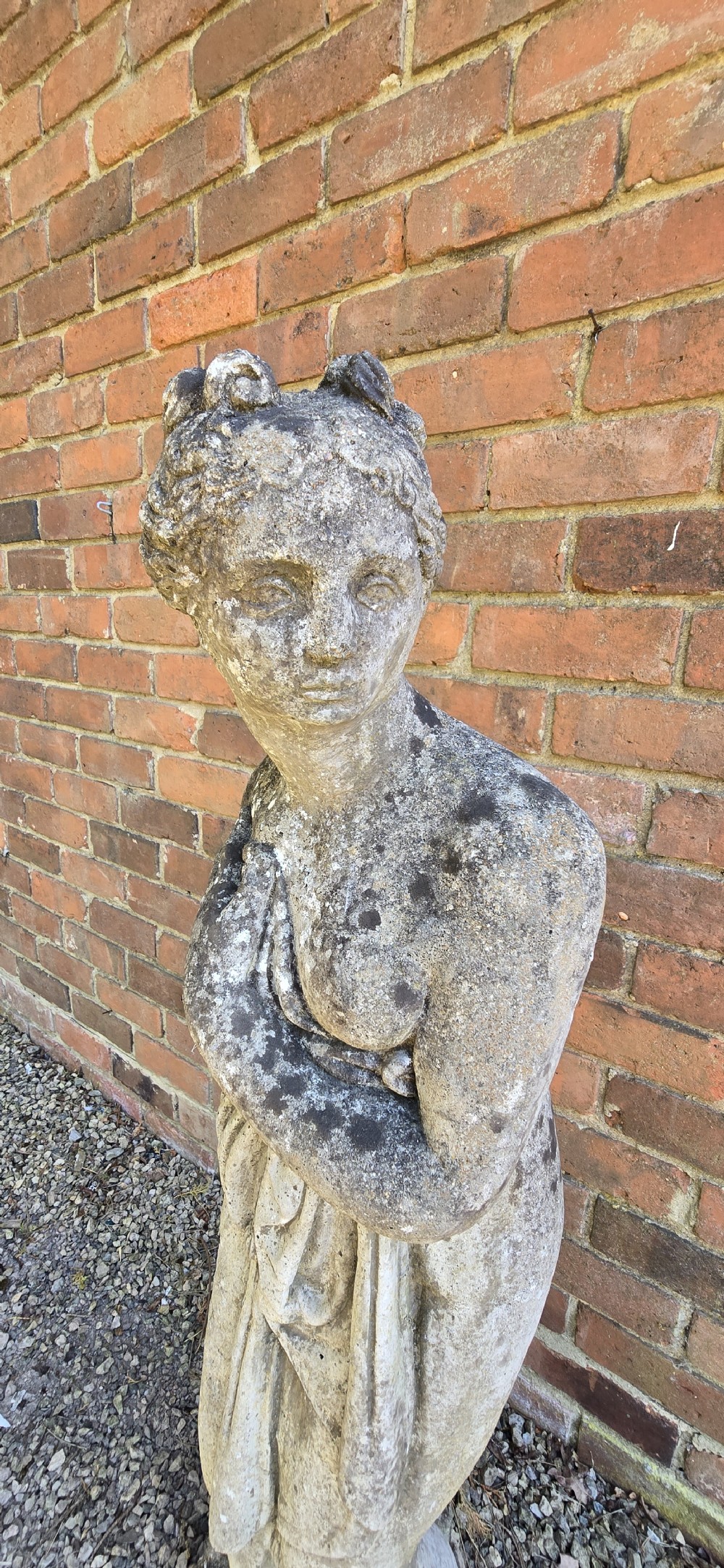 A weathered cast stone garden statue depicting a classical female figure draped in fabric standing on a round integral base. View VI