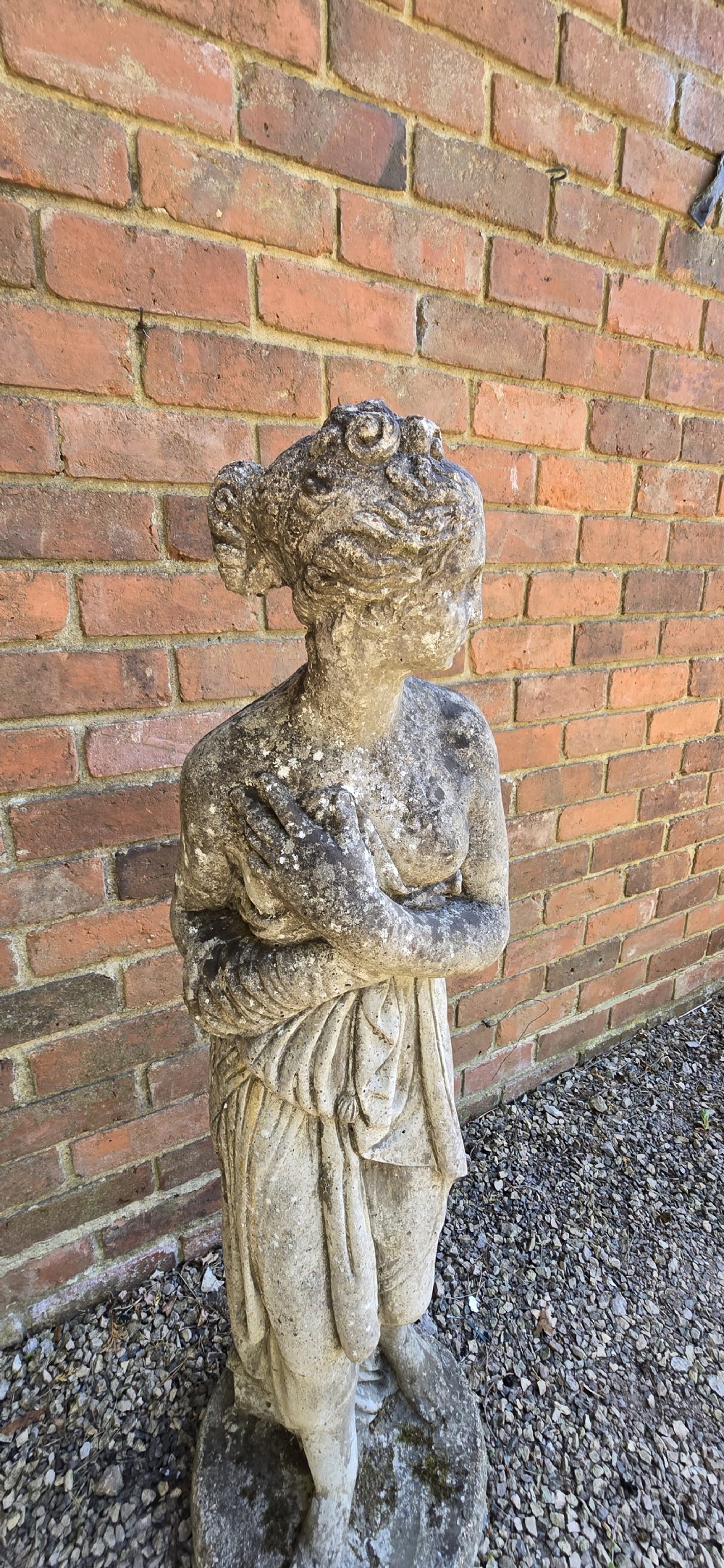 A weathered cast stone garden statue depicting a classical female figure draped in fabric standing on a round integral base. View VII