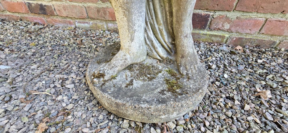A weathered cast stone garden statue depicting a classical female figure draped in fabric standing on a round integral base. View VIII