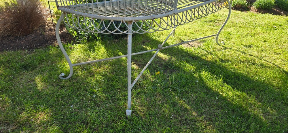 A weathered off-white wrought iron garden love seat with an S-shaped double seat and vertical wirework backrest. View V
