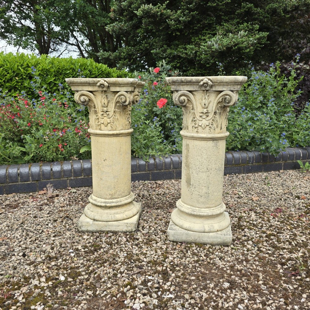 A pair of weathered buff cast stone Corinthian columns featuring acanthus leaf capitals and square bases.