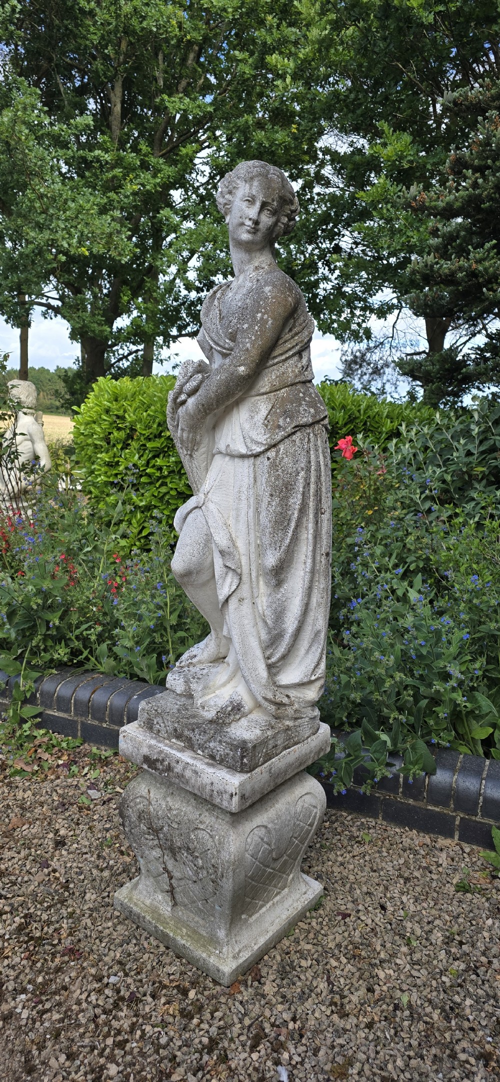 A full-length cast stone statue of a classical draped female figure standing atop a square carved plinth base. View III